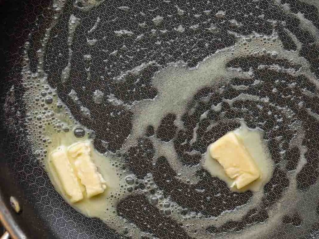 Two pieces of butter melting in a frying pan, creating swirls of melted butter and bubbles on the pans surface.